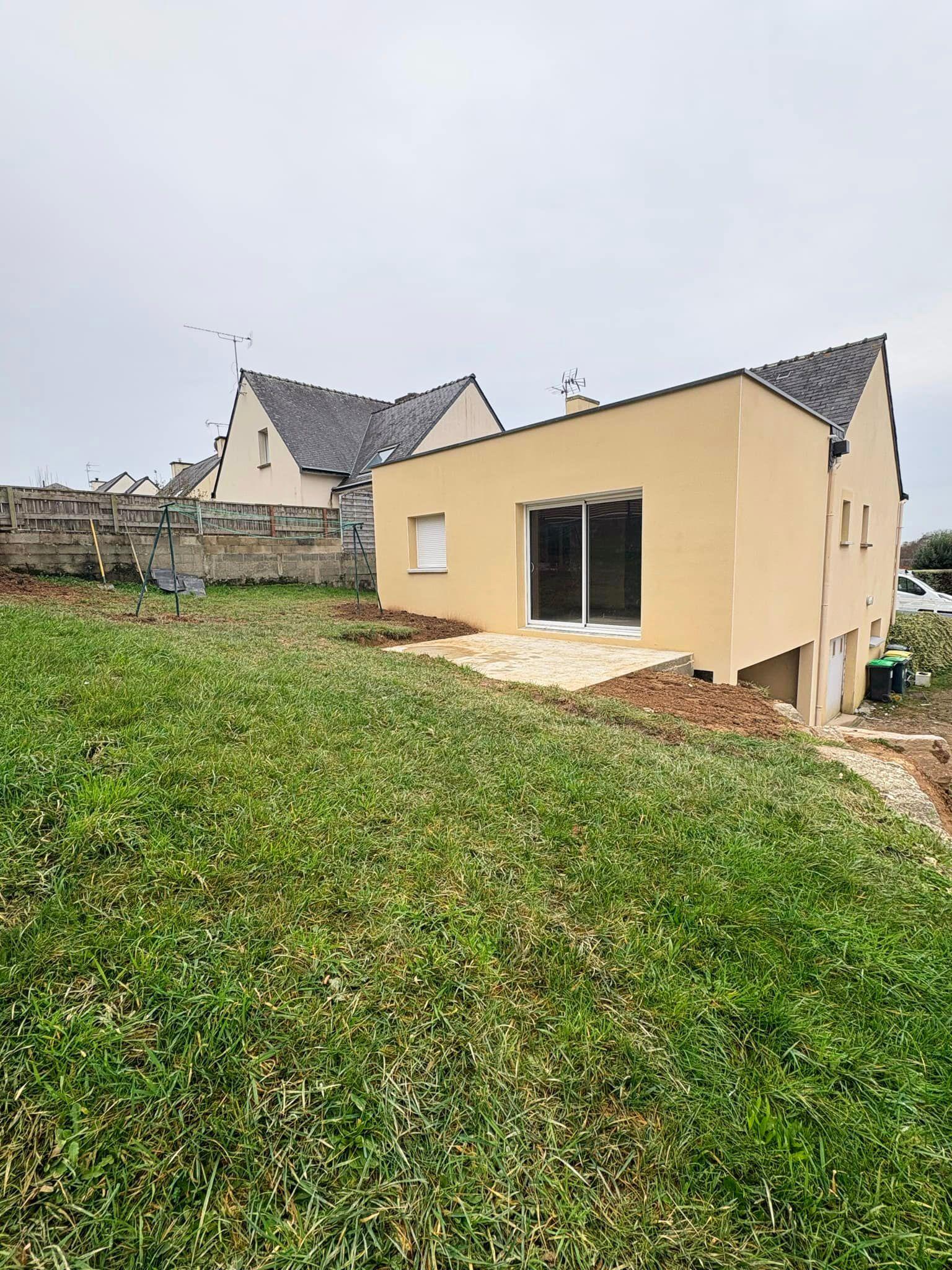 Extension de maison en agglos enduit ton clair beige, avec menuiseries alu et pvc blanc, toit plat en EPDM étanchéité, vue extérieure avec la terrasse en dallage brut.